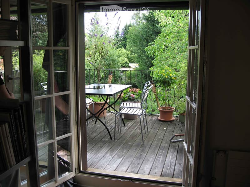 Balcony with wooden decking, glass sliding doors, table, chairs, and potted plants.