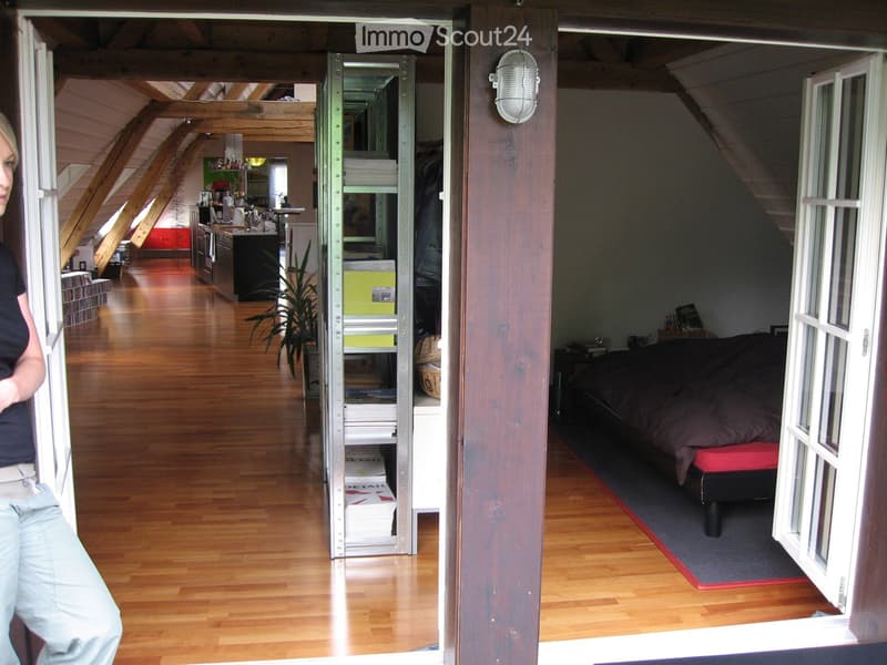 Open-plan attic space with wooden floors, featuring a bed, kitchen area, and bookshelf. Visible objects include a bed, kitchen counter, fridge, bookshelf, plant, and lamp.