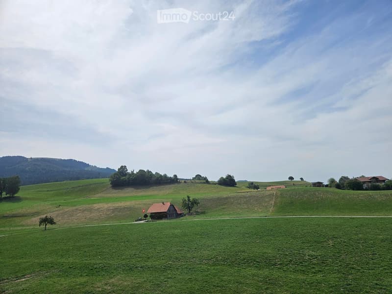 A peaceful rural landscape with rolling green hills, scattered trees, and a small wooden house with a red roof. The area is open and expansive, with a clear sky and distant mountains visible.