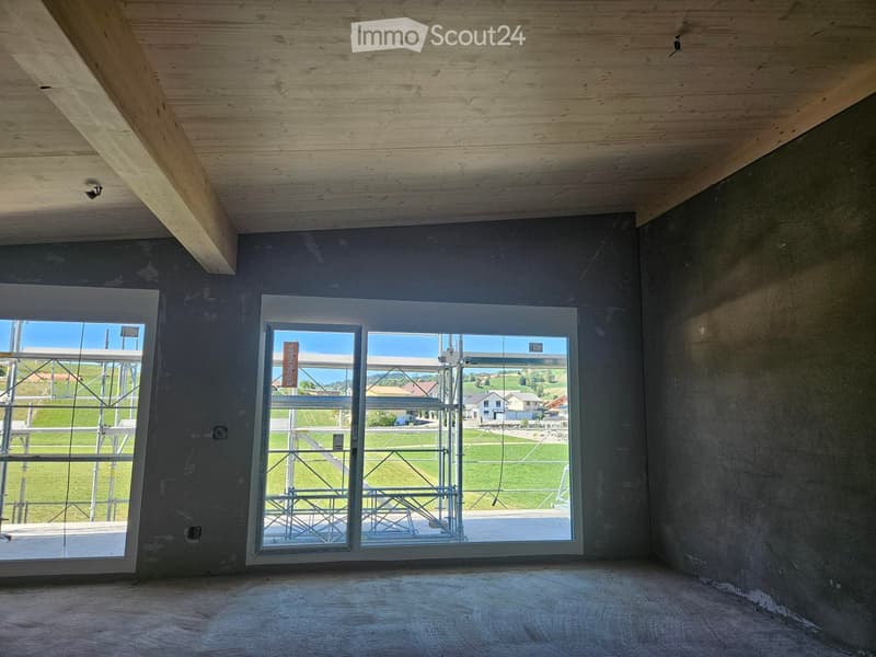Empty room with unfinished concrete walls and wooden ceiling beams, large windows showing outdoor view.