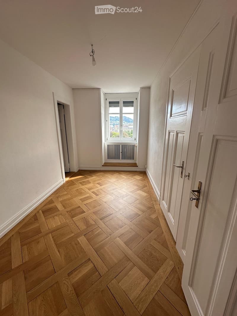 Empty hallway with wooden herringbone flooring, white walls, and several doors. A window with a radiator below it is visible at the end of the hallway.