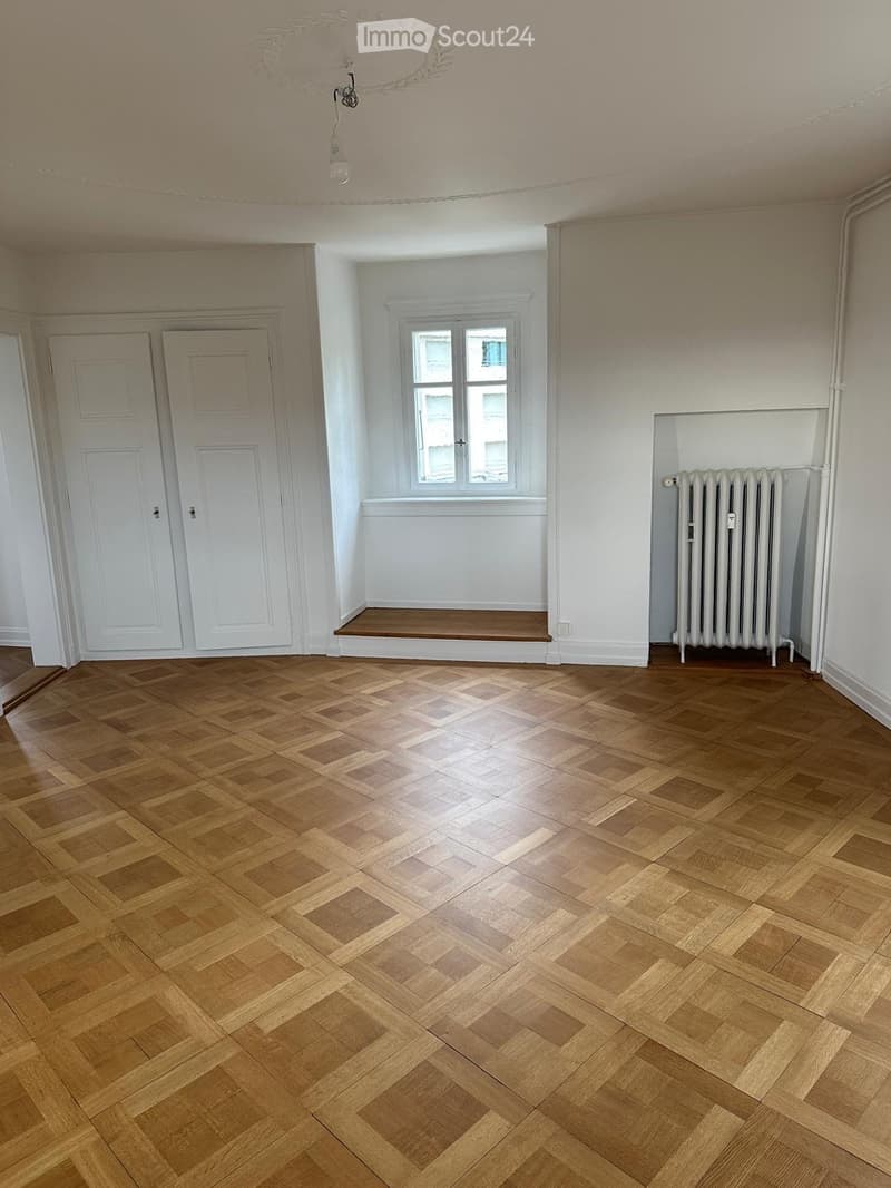 Empty room with wooden herringbone flooring, white walls, two white doors, a window with a wooden sill, and a radiator.