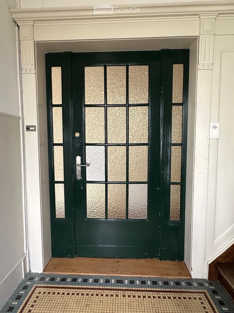 A hallway with a green double door featuring frosted glass panels, a wooden threshold, and a patterned tile floor.