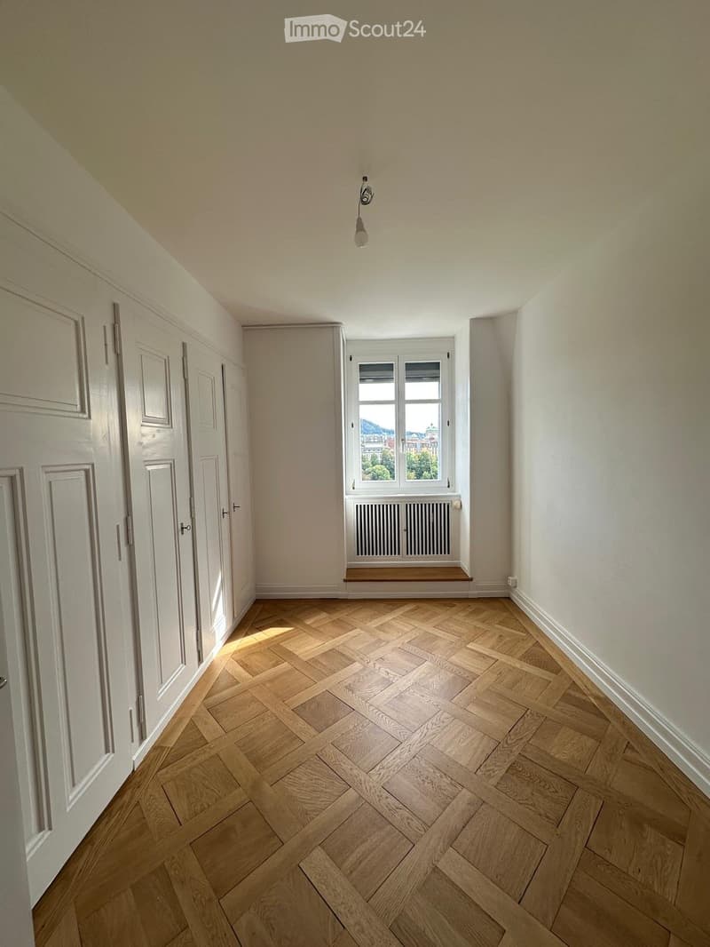 Empty room with wooden herringbone flooring, white walls, and a window with a radiator below. The room has several white doors on the left side and a single light fixture hanging from the ceiling.