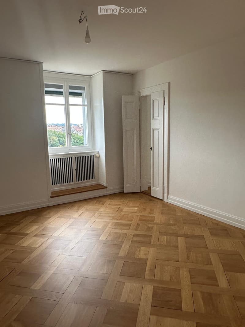 Empty room with wooden herringbone flooring, white walls, large window with radiator, and a single door.