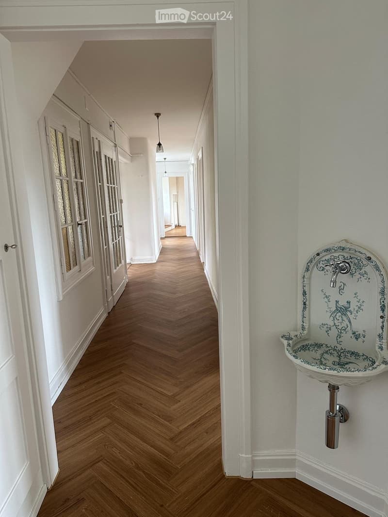 Elegant hallway with herringbone wooden flooring, white walls, and a decorative wall-mounted sink with a bird motif on the right side.