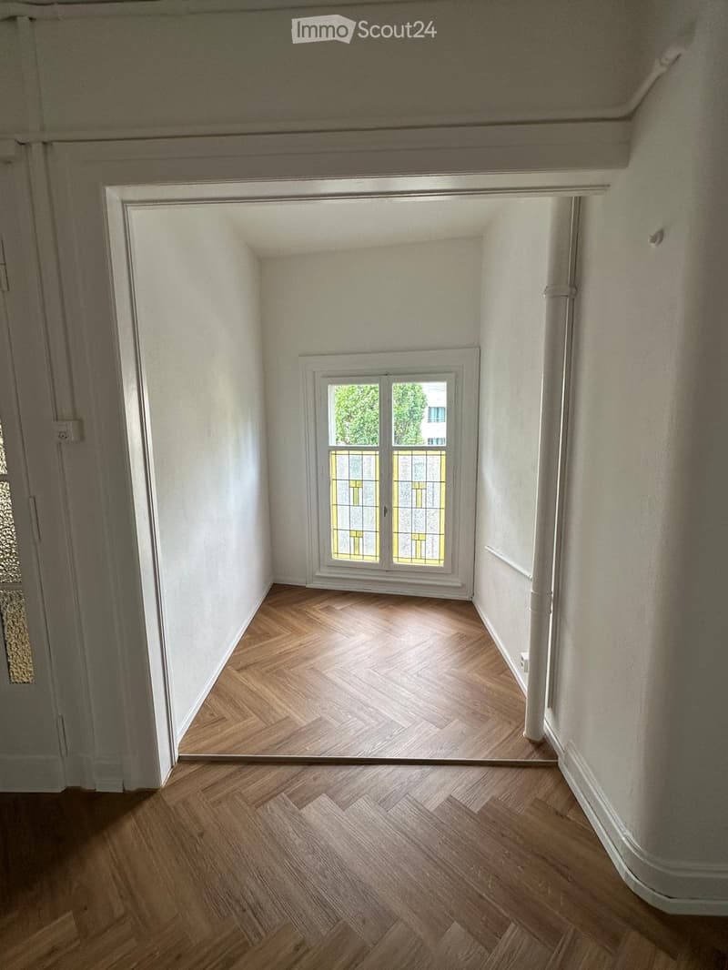 Empty room with wooden herringbone flooring, white walls, and a window with decorative glass panes.