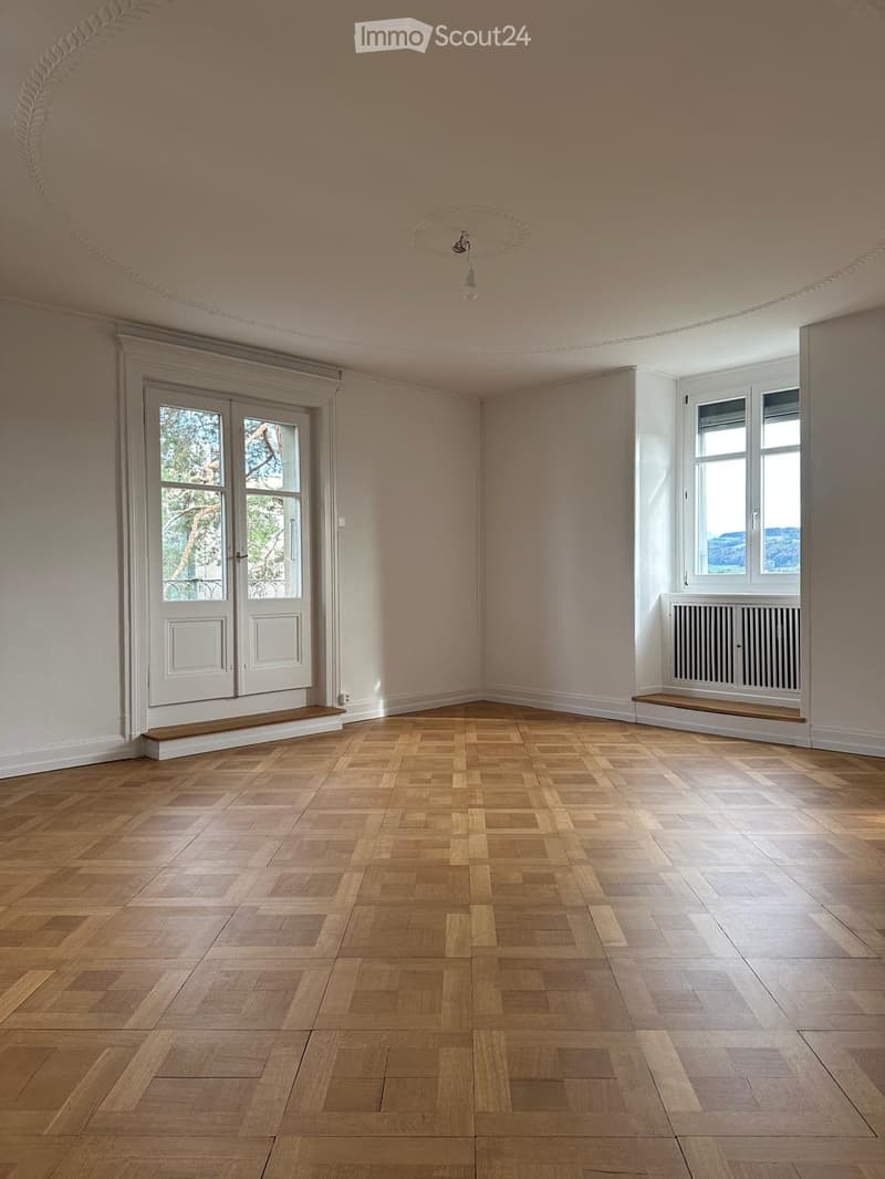 Empty room with wooden parquet flooring, light-colored walls, large windows, and a double door leading outside.