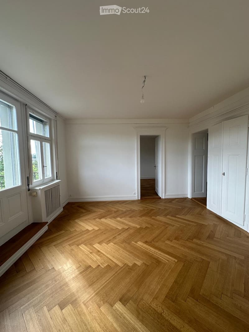 Empty room with wooden herringbone flooring, white walls, large windows, and a radiator. No furniture or equipment present.
