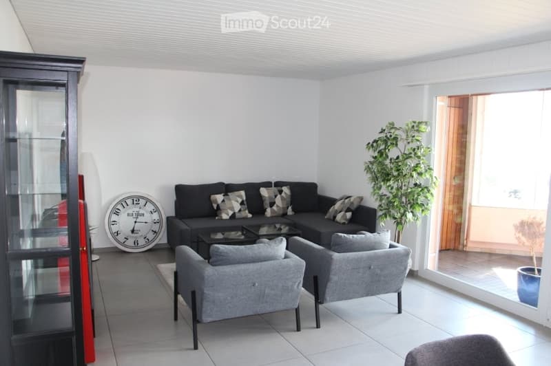 Modern living room with white walls, grey sofa, two armchairs, coffee table, large clock, cabinet, and a potted plant near the balcony door.