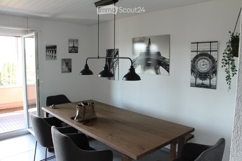 Dining room with a wooden table, four chairs, a handbag on the table, and several picture frames and a clock on the wall. A pendant light fixture hangs above the table.