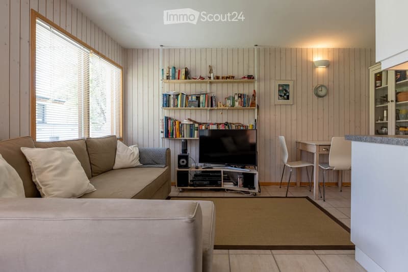 Furnished Room. Cozy living room with wooden panel walls, a large window with blinds, a beige sofa with pillows, an ottoman, a TV stand with a television, a bookshelf filled with books, a small table with chairs, and a wall-mounted clock.