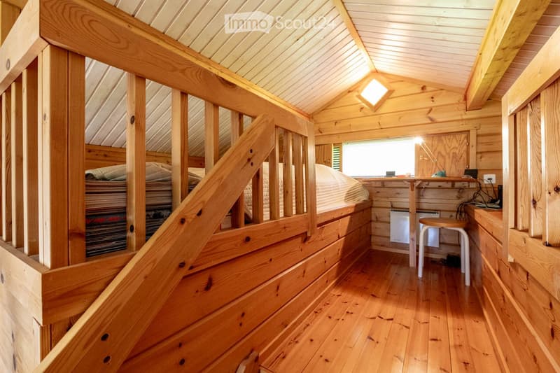Furnished Room. Cozy wooden loft bedroom with a raised bed, built-in desk, and small stool. Features wooden walls, slanted ceiling, and warm lighting.