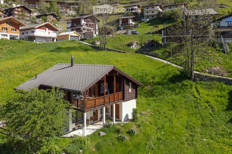 House. A two-story wooden house with a balcony and a small patio, surrounded by green hills and other houses in a rural setting.