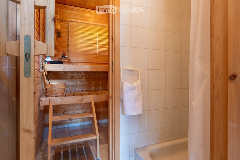Bathroom. Bathroom with a wooden sauna area featuring a wooden bench, bucket, and ladder, adjacent to a tiled bathroom area with a bathtub and a towel hanging on a rack.