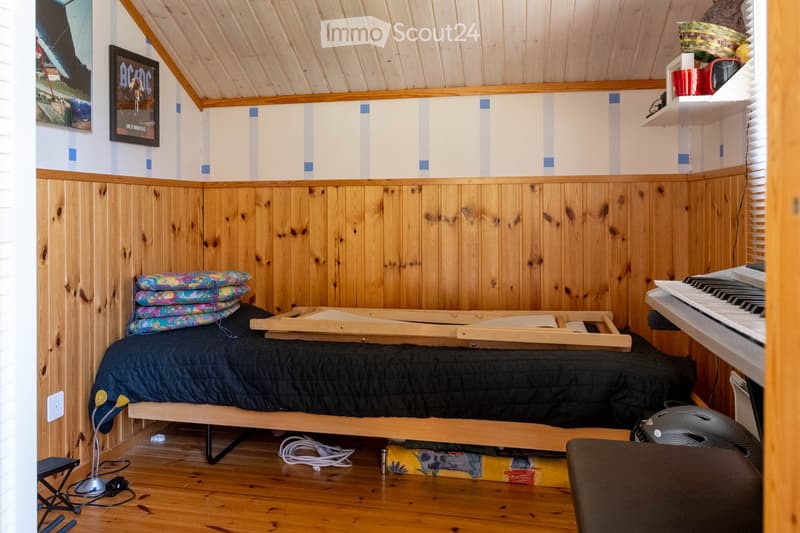 Furnished Room. Cozy attic bedroom with wooden walls and ceiling, featuring a bed, small table, piano, shelves, and a picture frame on the wall.
