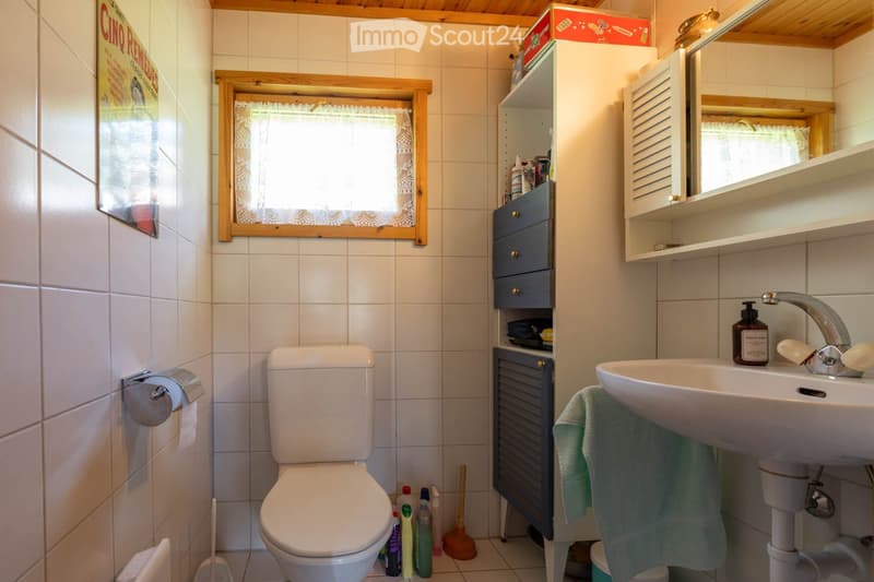 Bathroom. A small bathroom with white tiled walls and floor, featuring a toilet, sink with a mirror, a small cabinet, and cleaning supplies on the floor.