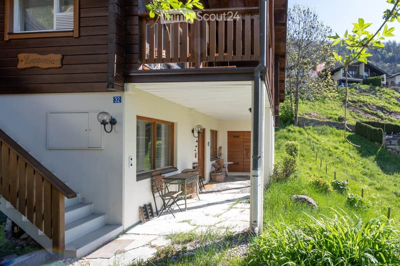 Building. Two-story wooden house with a balcony, outdoor seating area, and lush green surroundings.