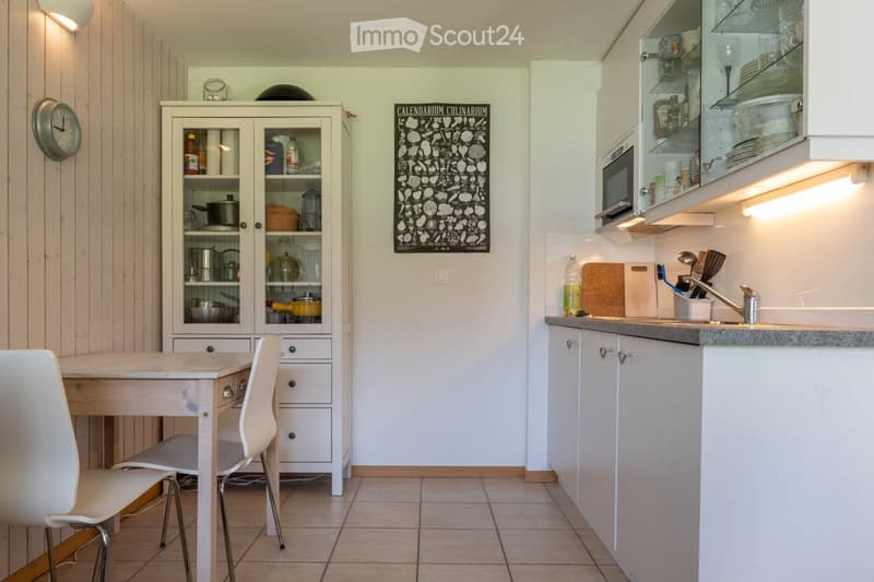 Kitchen. The kitchen features a white table with a single chair, a white cabinet with glass doors displaying dishes and cookware, a microwave, a sink with a faucet, and a cutting board on the counter.