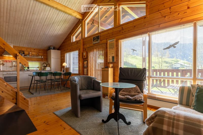 Furnished Room. Cozy living room with wooden walls, large windows, and a kitchen area. Features a sofa, chairs, a coffee table, and a dining area with stools and a table.