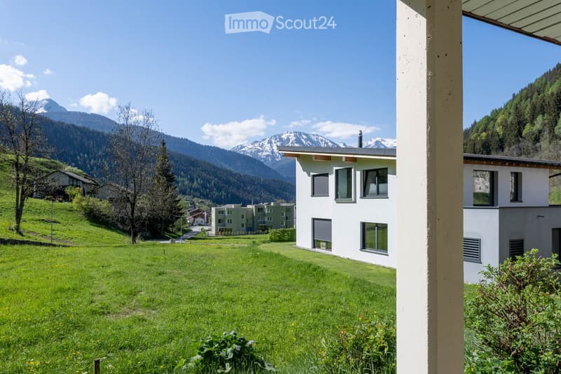 Building. Modern white building with large windows, surrounded by green lawn and trees, with snow-capped mountains in the background.