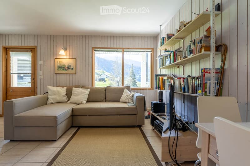 Living Room. A cozy living room with a sectional sofa, a small table, a chair, shelves filled with books, and a television. The room features wooden panel walls, a large window with a scenic view, and a warm, inviting atmosphere.