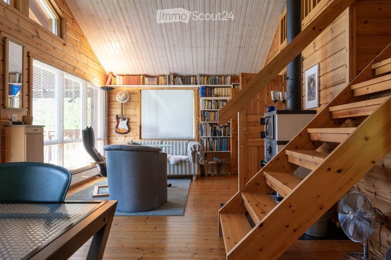 Furnished Room. Cozy living room with wooden walls and ceiling, large windows, a staircase leading to an upper level, a sofa, a round stool, a table with chairs, a bookshelf, and a guitar on the wall.