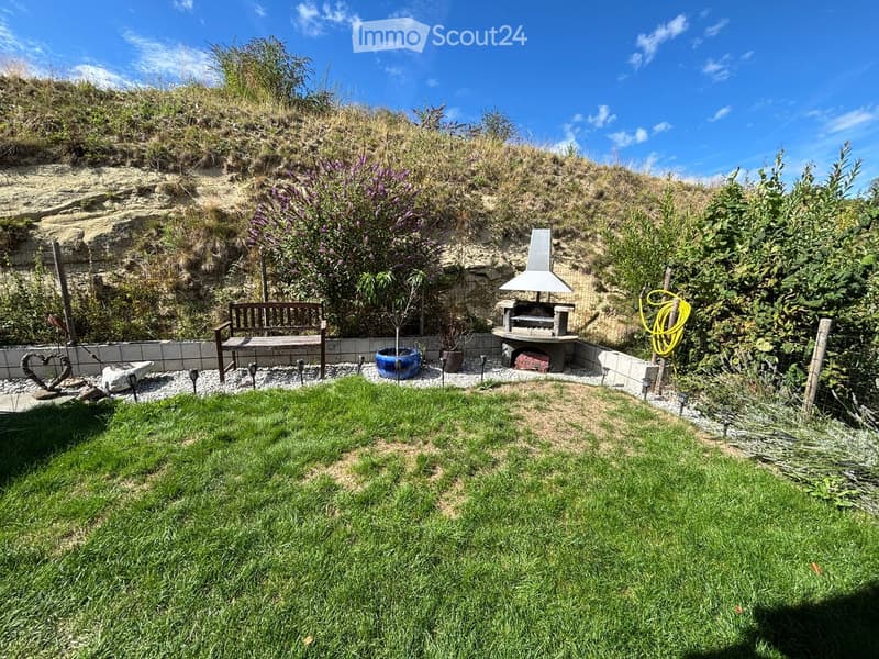 Well-maintained garden with a wooden bench, barbecue, potted plants, and garden lights, surrounded by greenery and a sandy hill.