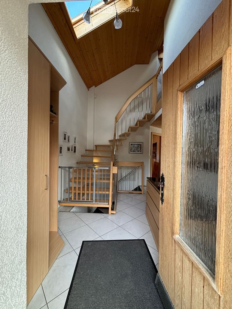 Spacious hallway with wooden staircase, metal railing, tiled floor, and a black runner. Features a wooden door, a small cabinet, and framed pictures on the wall.