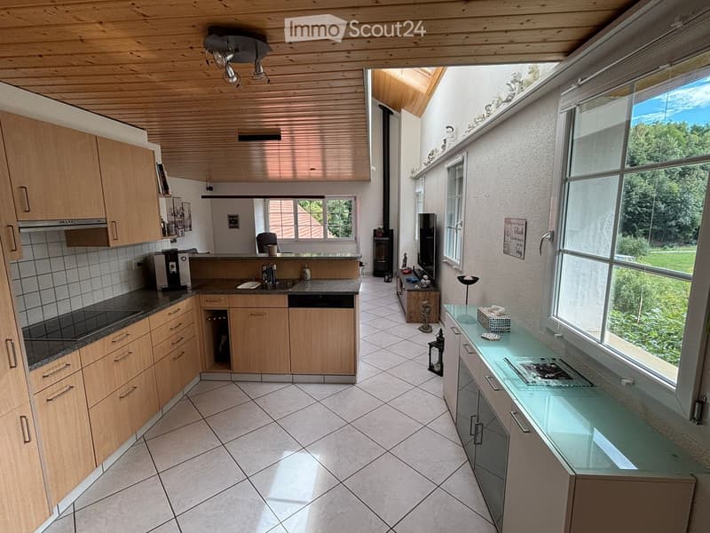 Bright kitchen with wooden cabinets, granite countertops, and a central island. Features include a sink, dishwasher, coffee maker, and a small seating area with a table and chair. Large windows provide natural light and a view of the outdoors.