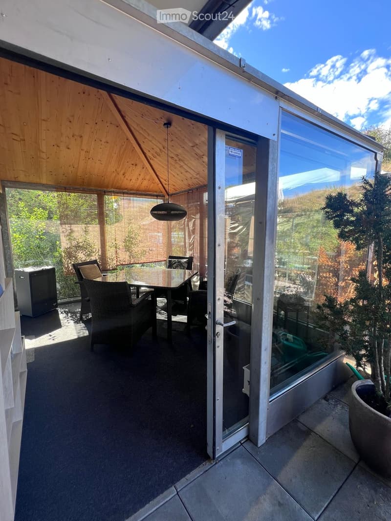 A modern, well-lit room with a glass door leading to an outdoor area, featuring a dining table, chairs, and a hanging lamp.