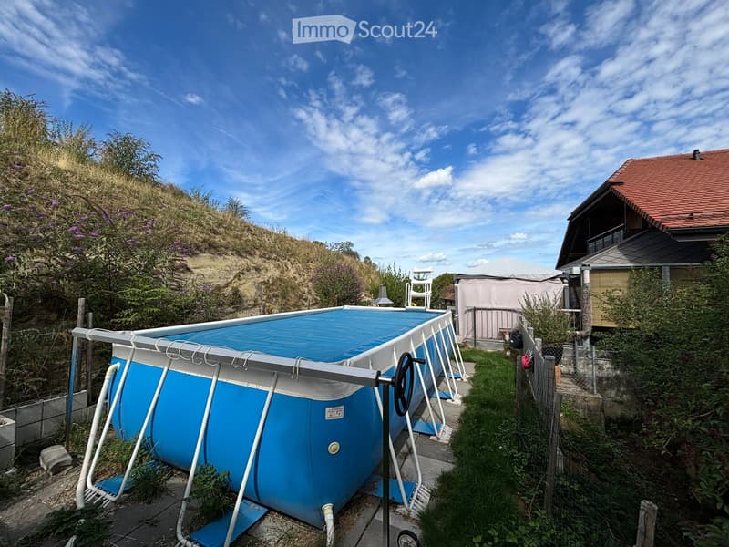 Outdoor area with a blue inflatable swimming pool, surrounded by greenery, a fence, and a nearby house with a red-tiled roof.
