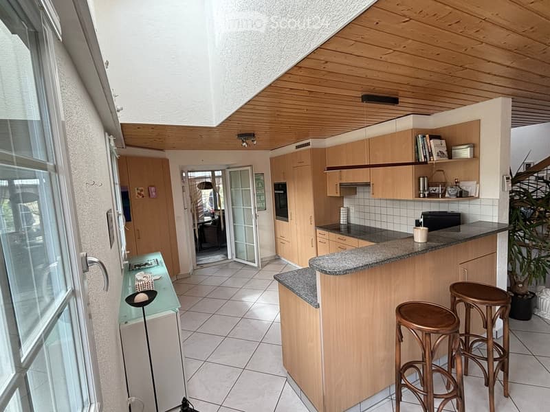 Bright kitchen with wooden cabinets, granite countertops, and a small seating area with two stools. Features an open layout with a glass door leading outside.