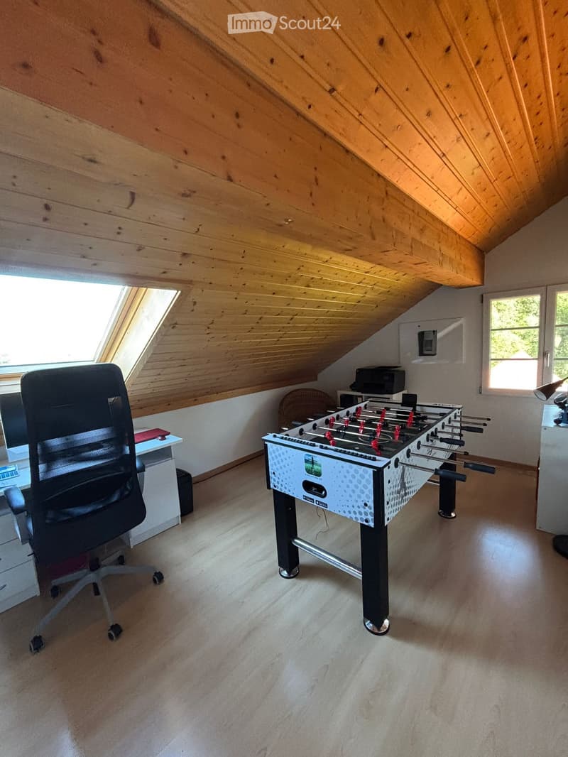 Bright attic room with wooden slanted ceilings, large windows, and skylight. Contains an office chair, a small table, a futon, and a foosball table.