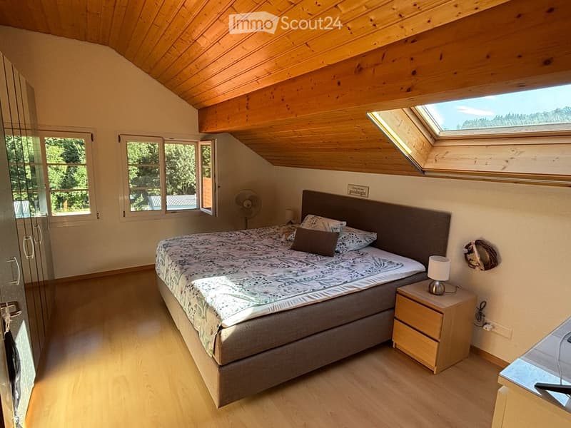 Cozy attic bedroom with wooden slanted ceilings, large windows, and skylights. Features a double bed with patterned bedding, a nightstand with a lamp, and a fan near the window.