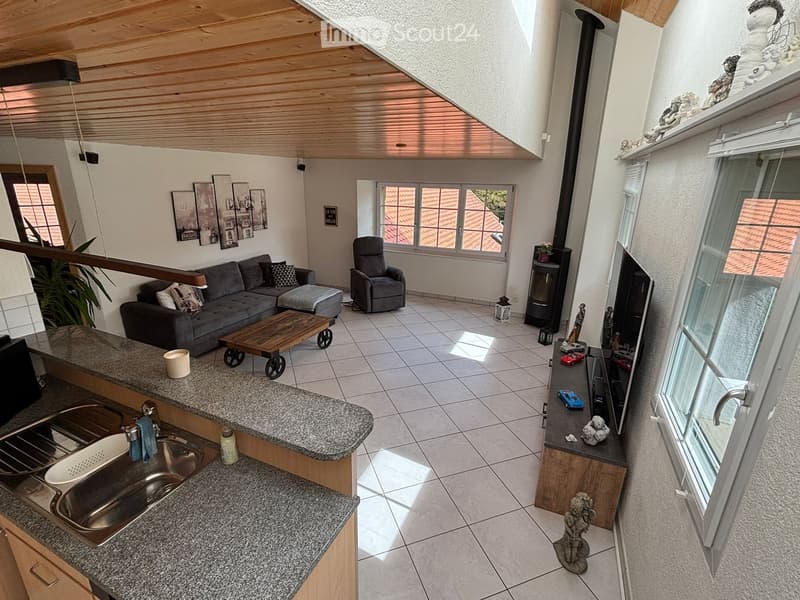 Open-plan living and dining area with a grey sofa, coffee table, armchair, sink, and fireplace. The space features large windows and a wooden ceiling.