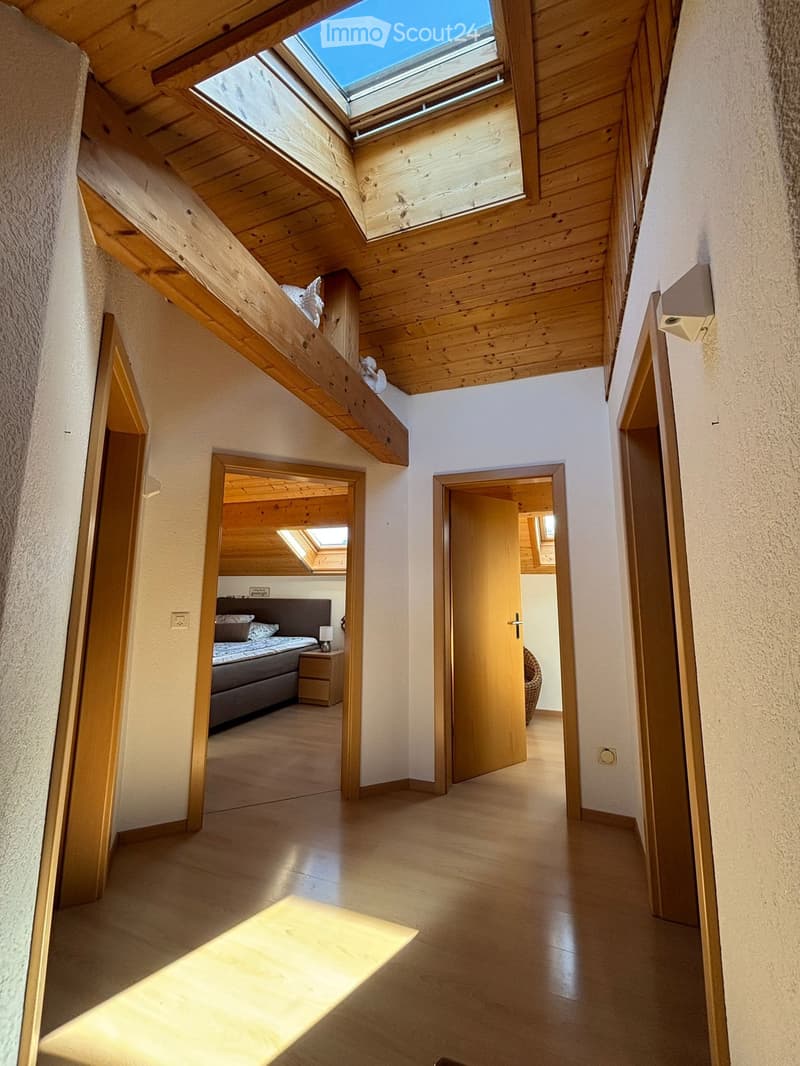 Bright hallway with wooden beams and skylight, leading to a bedroom with a bed, nightstand, and lamp.