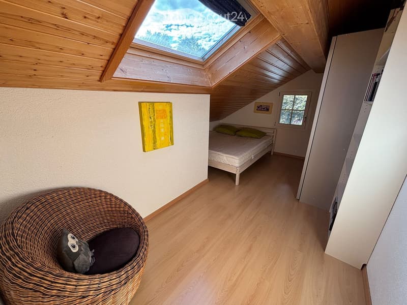 Cozy attic bedroom with wooden floors, slanted wooden ceiling, skylight, bed with pillows, wicker chair, and artwork on the wall.