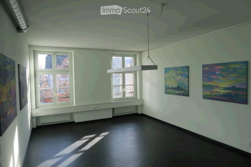 Empty room with bare walls, wooden floor, large windows, and several paintings on the walls.