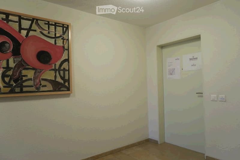 Empty room with light-colored walls, wooden floor, and a single door. A framed abstract painting is mounted on one wall.