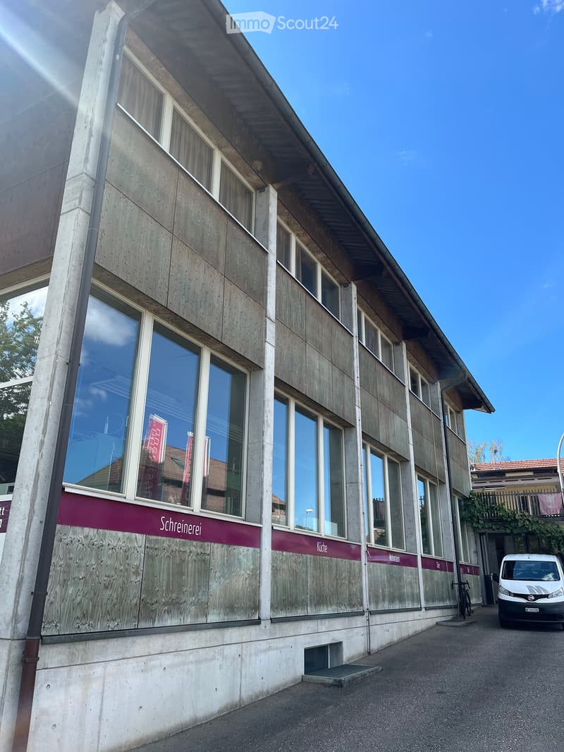 Modern two-story building with large windows and wooden paneling, featuring signage for "Schreinerei" and "Küche" on the facade.