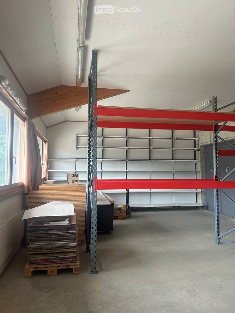 An empty, unfinished room with exposed wooden beams, metal shelving units, and a concrete floor. The room has large windows with curtains and a stack of materials on a pallet.