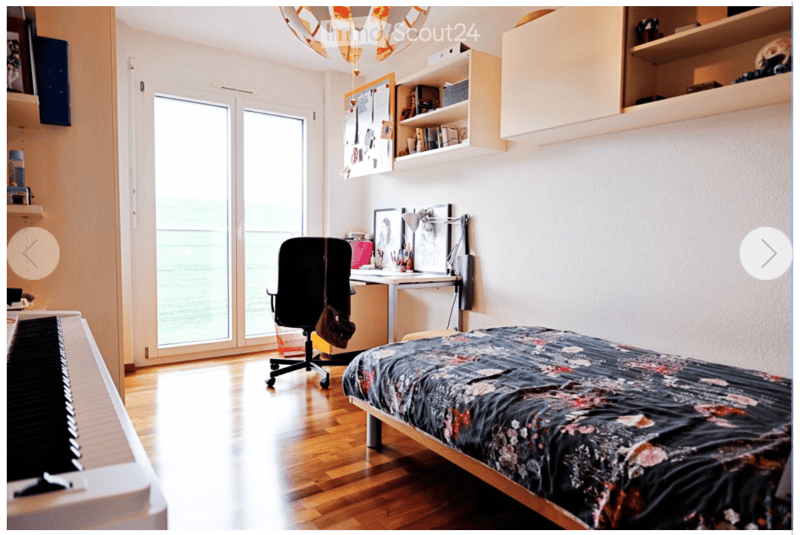 Bright bedroom with wooden flooring, large window offering a view, bed with patterned bedding, desk with chair, shelves, and a piano.