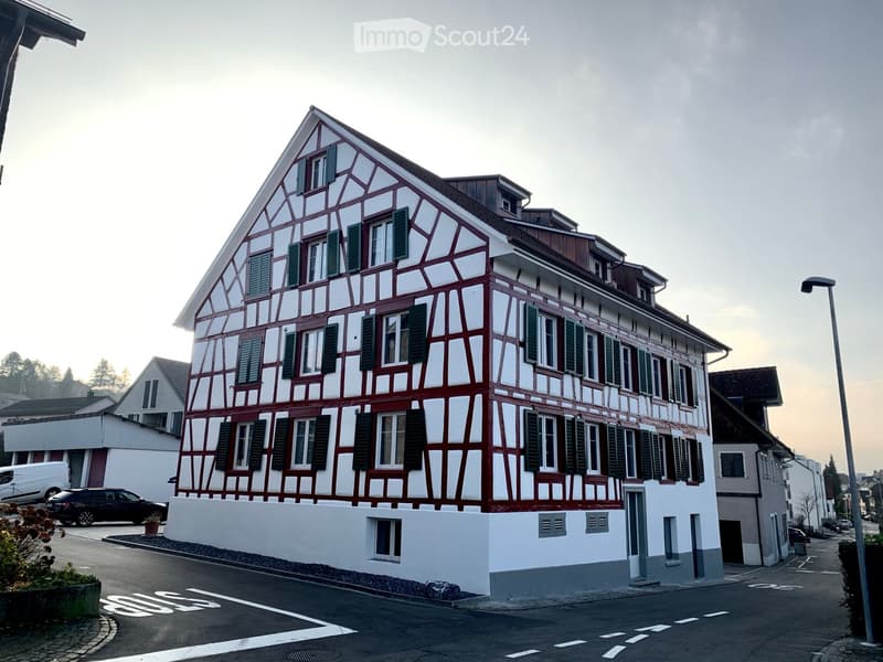 A traditional Swiss half-timbered house with white and red painted walls, green shutters, and multiple windows, situated on a street with a low white wall in front.
