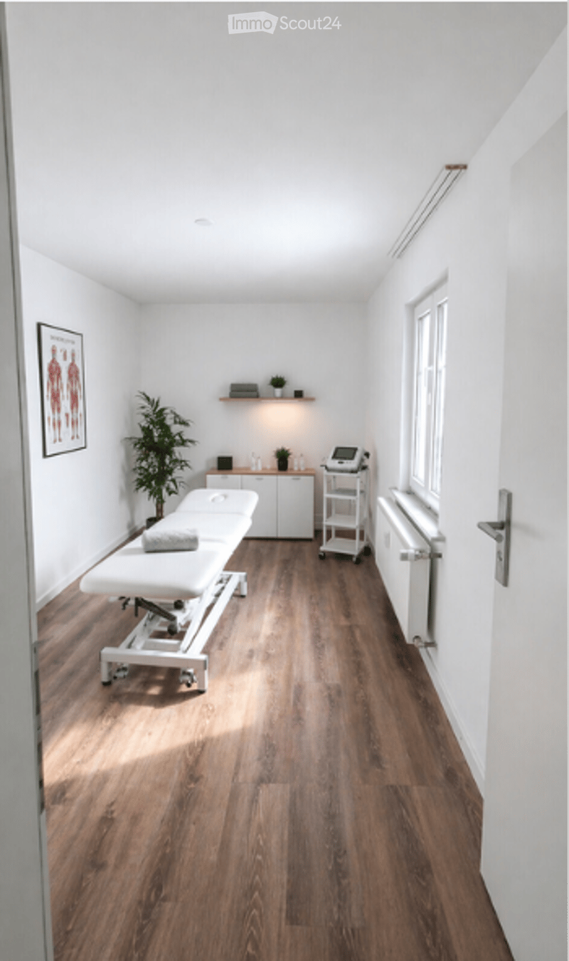 A clean, modern treatment room with a wooden floor, white walls, a treatment table, a small cabinet, a potted plant, and a radiator.