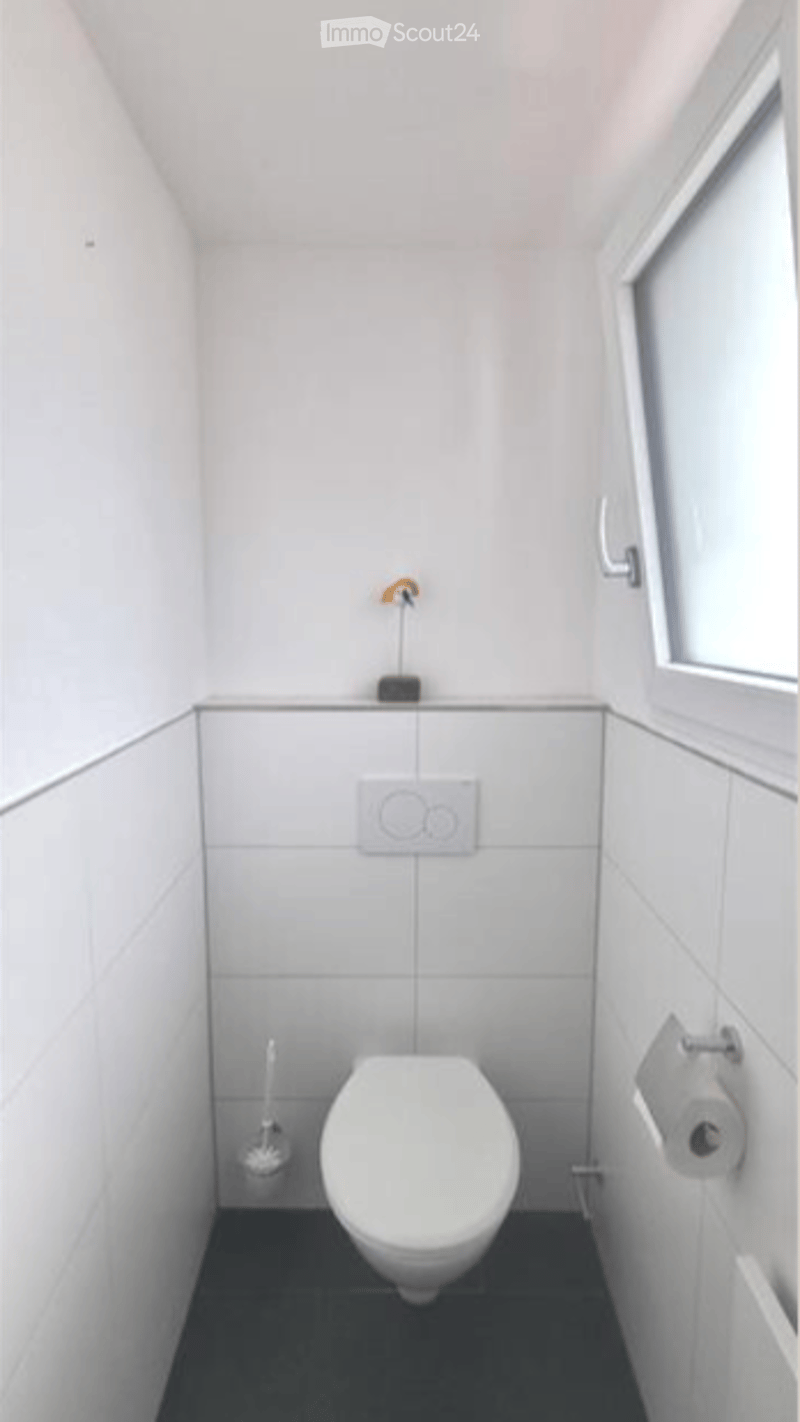 Modern, compact bathroom with white tiled walls and floor, a toilet, toilet paper holder, toilet brush, and a light switch.