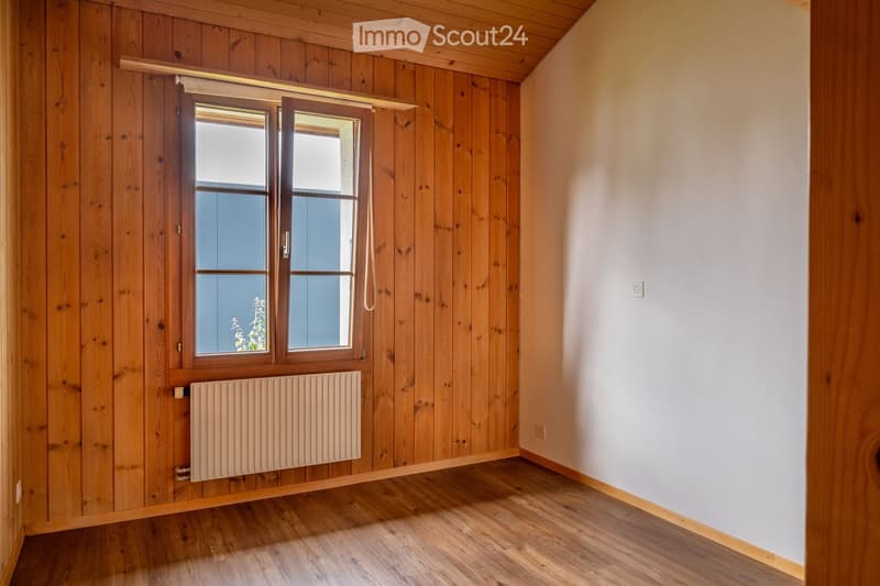 Empty room with wooden walls and flooring, featuring a large window with wooden frame and a radiator below it.
