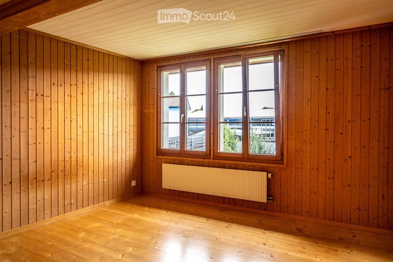 Empty room with wooden walls and floor, large window with wooden frame, radiator below the window.