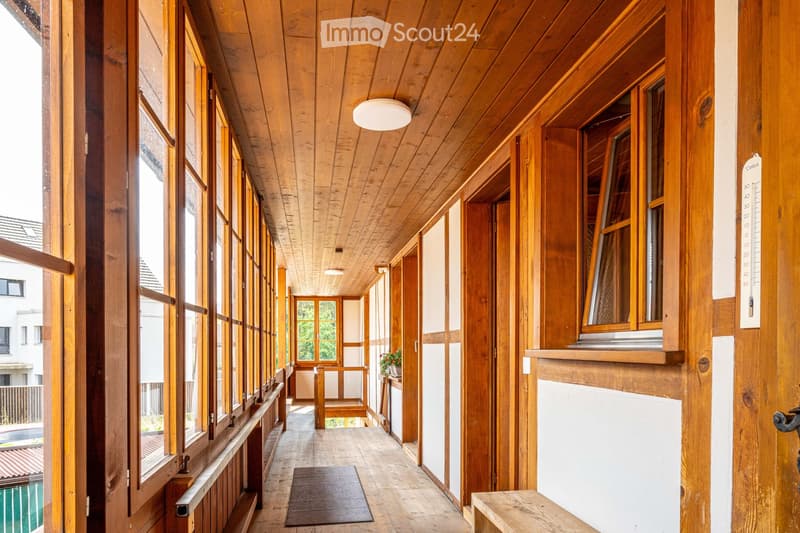 Wooden hallway with large windows, wooden ceiling, and a small bench at the end.