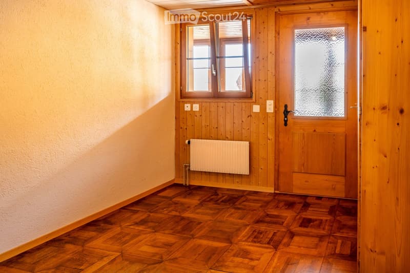 Empty hallway with wooden flooring, light-colored walls, a window with blinds, a radiator, and a wooden door with a glass panel.